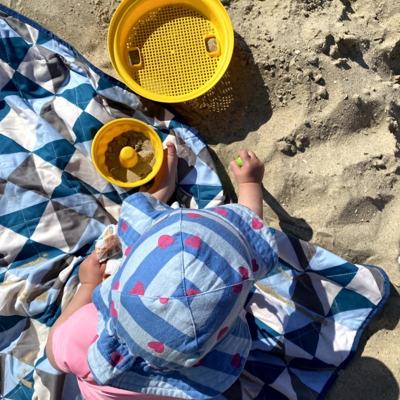 Kind am Strand, Sicht aus der Vogelperspektive auf Kind mit Mütze, das auf einem Handtuch am Strand sitzt mit gelben Sandförmchen, Urlaub